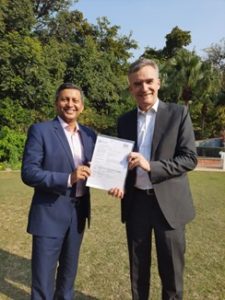 Pearson India and Chevening representative hold up a piece of paper that acknowledges the partnership agreed between the two organisations. Both men wear suits and stand in a park. 