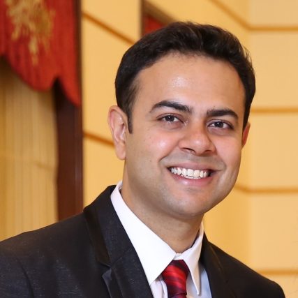 SUYASH MEHROTRA smiles at the camera, in a suit and red tie