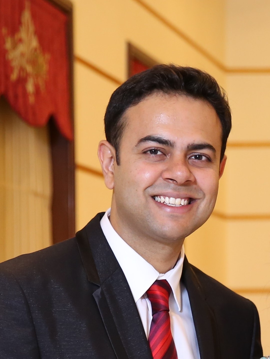 SUYASH MEHROTRA smiles at the camera, in a suit and red tie