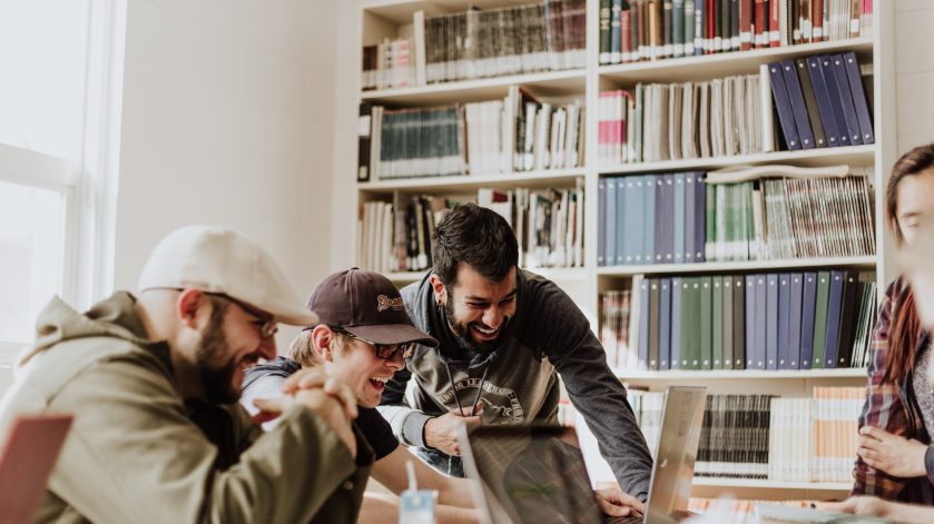 A group of friends joke together whilst in group study. The 3 young men sit around a table with their laptops in front of a bookshelf.