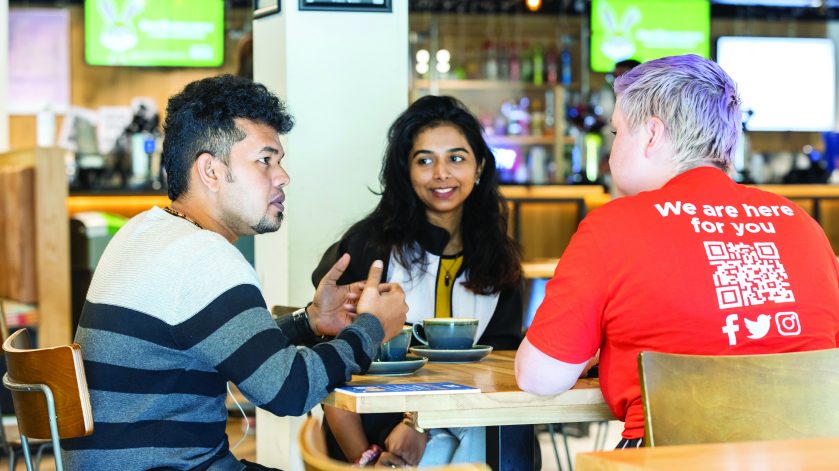 Two students sitting at a table talking to a support staff member.