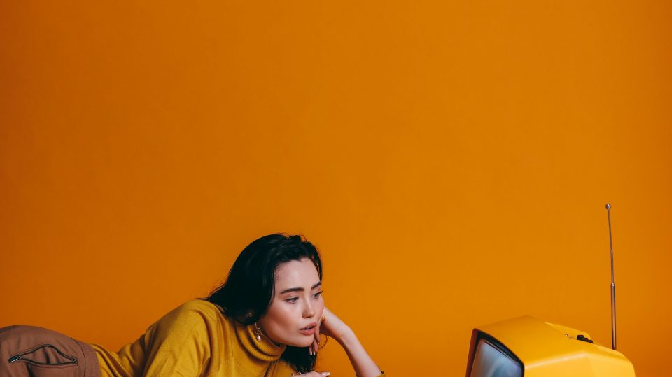 A woman lying on a bed with bright orange wall behind her and she is looking at an old fashioned, 1960s TV