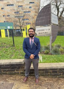 Kayan stands in front of a University of Bradford sign.