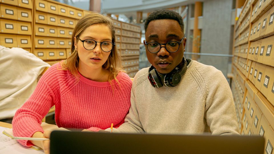 A man and a woman sat in a room with lockers, with papers on a desk and both looking at a laptop.
