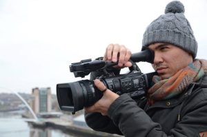 Person wearing a gray knit hat and scarf filming outdoors with a professional video camera near a river and cityscape.