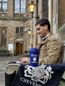 Scholar sits on a bench on the university grounds with a Chevening branded tote bag and coffee cup. 