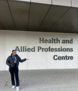 Scholar stands in front of a building that says 'Health and Allied Professions Centre'. 
