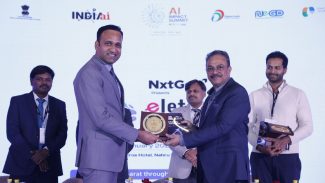 Two men in suits exchanging an award plaque on stage at the AI Impact Summit event with other attendees in the background