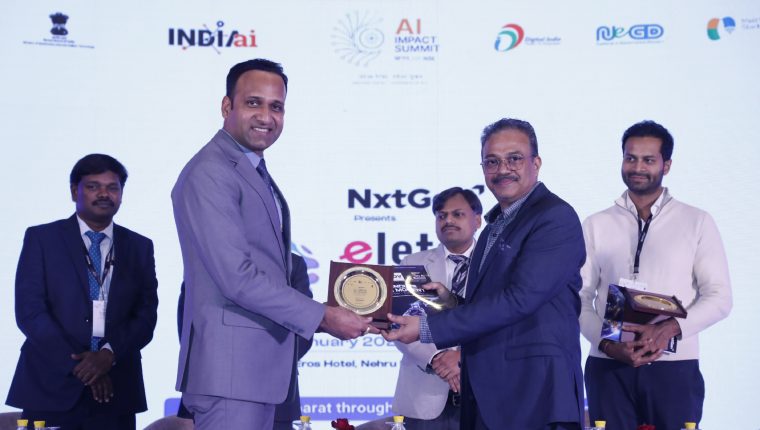 Two men in suits exchanging an award plaque on stage at the AI Impact Summit event with other attendees in the background